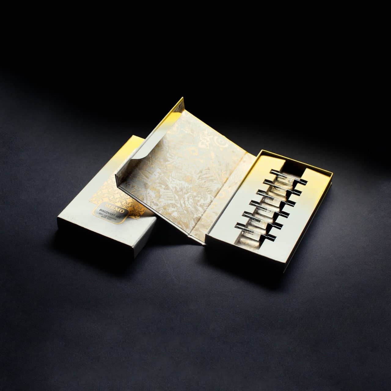 Elegant gold and white gift box containing seven sleek, metallic travel-sized bottles arranged in a row.