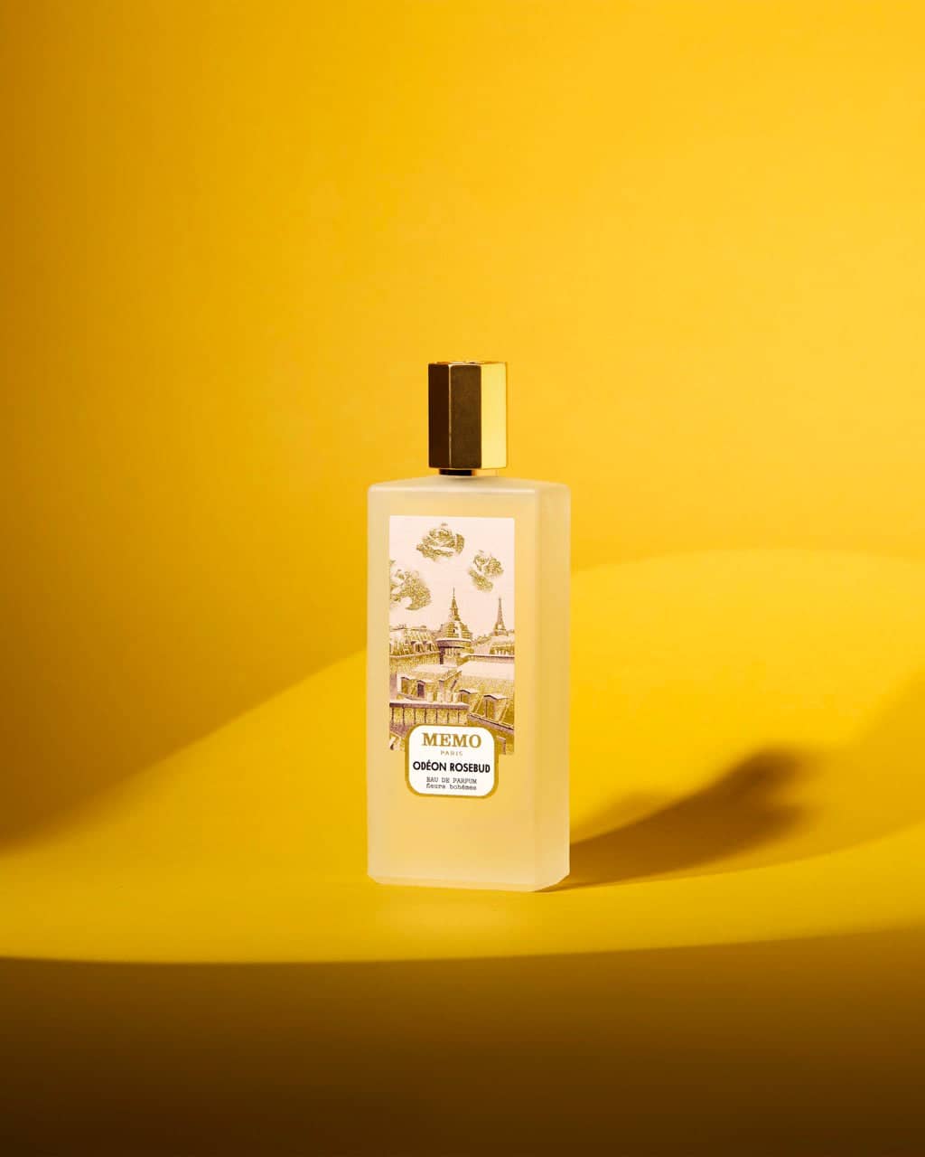 A translucent, rectangular bottle of memo paris perfume with a gold cap and an illustrated label depicting architectural scenes.